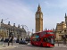 Red double decker bus driving near Big Ben and the Houses of Parliament in central London on a clear day, with the London Eye visible in the background.
