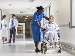 A nurse gently talks to a patient in a wheelchair in a bright hospital corridor, while two doctors chat in the background.