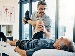 A male physiotherapist helps an older man stretch his leg during a therapy session, with anatomical charts and natural light in the background.