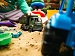 Close-up of a child playing in a sandpit with toy dump trucks, buckets, and moulds; one green truck in focus and a blue truck blurred in the foreground.