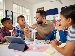 Teacher sitting at a classroom table with a mixed group of children, explaining a science activity that uses small model wind turbines and a digital tablet, as the pupils listen and discuss their ideas together in a bright, modern classroom.