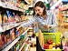 A smiling woman shopping in a supermarket aisle with a trolley full of groceries, symbolising Asda’s trial of AI-powered cameras that scan shelves to detect empty spaces and expired products.