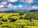 A bright rural South Wales landscape with a patchwork of green fields, hedgerows, scattered farmhouses and woodland, leading to low hills under a blue sky filled with white clouds.
