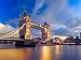 A wide-angle view of London’s Tower Bridge and The Shard at dusk, with the River Thames in the foreground reflecting the city lights under a dramatic sky.