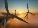 Wind turbines standing tall above the clouds at sunrise, with blades turning in golden light, symbolising renewable energy and modern technology.