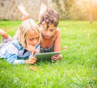 Two children lying in the grass and studying a computer tablet